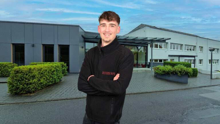 Mann in schwarzer Jacke mit verschränkten Armen vor einem modernen Firmengebäude; im Hintergrund Eingangsbereich, Grünpflanzen und blauer Himmel.